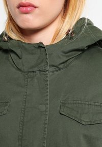 Chaqueta verde oliva con capucha, con dos bolsillos en el pecho, cierre de cremallera y herrajes decorativos en la capucha. Textura de tela suave.