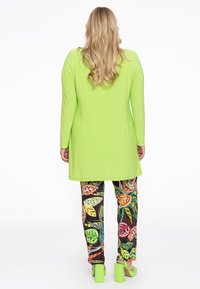Heldere groene lange mouw blouse in combinatie met kleurrijke, gemusterde broek met bladdessin op een donkere achtergrond en groene blokhak schoenen.