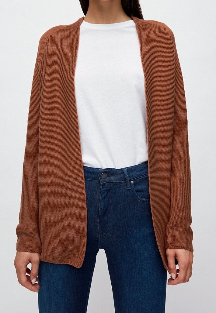 Femme portant un cardigan en maille marron ouvert sur l’avant, par-dessus une chemise blanche et un jean bleu, vue des épaules aux hanches, sur un fond uni.