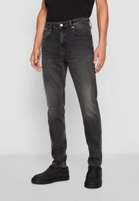 Vaqueros ajustados de color gris oscuro con un acabado desgastado, diseño de cinco bolsillos, remaches de metal y cierre clásico de botón. Combinados con zapatos negros.