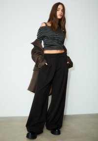 Schwarze weit geschnittene Hosen mit Falten, kombiniert mit einem grau gestreiften, schulterfreien Oberteil. Ein brauner übergroßer Mantel hängt über dem Arm. Schwarze Plateauschuhe.