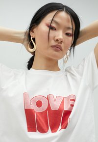Camiseta blanca con una gran impresión en degradado rojo que dice "AMOR" en el frente, combinada con pendientes en forma de lágrima dorados. Textura de tela suave.