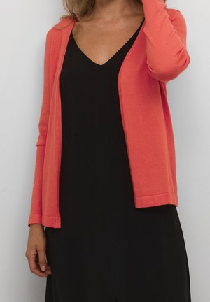 Femme portant un cardigan corail à manches longues sur une robe noire à col en V, debout devant un fond gris clair uni.