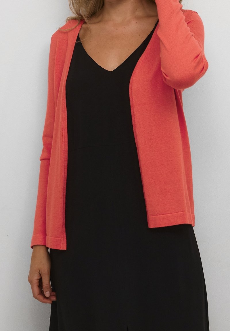 Femme portant un cardigan corail à manches longues sur une robe noire à col en V, debout devant un fond gris clair uni.