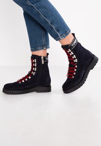 Námořnické semišové kotníkové boty s červenými tkaničkami, stříbrnými prvky a černými robustními podrážkami. Boční zip a logo na horní části.