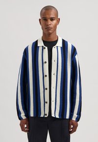 Neulottu neuletakki, jossa pystysuorat raidat sinisen, valkoisen ja mustan sävyissä. Edessä napitettu, kontrastivärinen kaulus. Löysä istuvuus ja teksturoidu kangas.