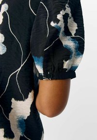 Top negro con tejido texturizado que presenta patrones abstractos en blanco y azul, mangas cortas abullonadas con detalles de botones y un corte relajado.