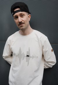 Cremefarbenes Langarmhemd mit einem schwarzen Schallwellen-Graphic und einem kleinen Logo-Patch am rechten Ärmel. Lässige Passform und weicher Stoff.