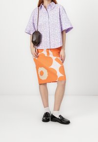 Chemise à manches courtes lavande avec imprimé floral, jupe crayon à motif orange, mocassins noirs avec accents décoratifs, et un petit sac marron.