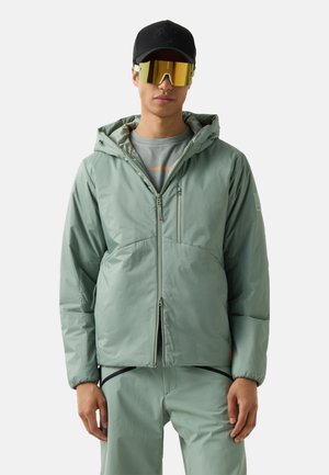 Man draagt een lichtgroene capuchonjas en bijpassende broek, een zwarte pet en grote reflecterende gouden sportzonnebril, staand tegen een witte achtergrond.