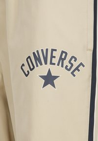 Beiga sweatpants med marinblå sidostreep och stor "CONVERSE"-text ovanför en stjärnlogotyp. Tillverkade av mjukt, slätt tyg.