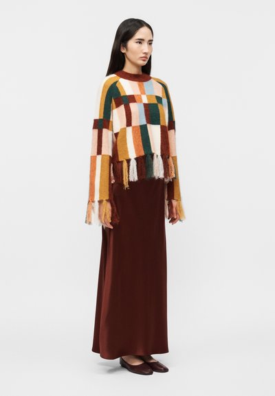 Femme portant un pull à carreaux multicolores avec des franges et une longue jupe marron, debout devant un fond clair uni.