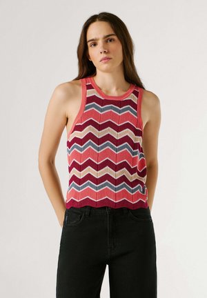 Femme portant un haut sans manches en tricot avec un motif en zigzag rouge, rose, beige et gris, associé à un pantalon noir, debout devant un fond uni.
