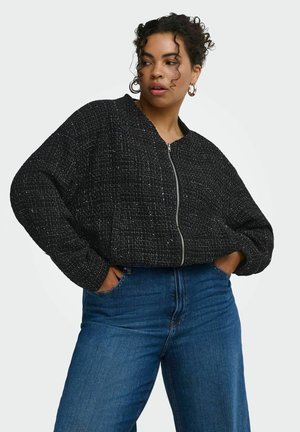 Femme aux cheveux bouclés, portant des boucles d'oreilles anneaux, une veste noire texturée zippée, et un jean bleu taille haute, les mains dans les poches.