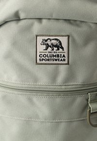 Το σακίδιο πλάτης Columbia Sportswear σε ανοιχτό πράσινο ύφασμα διαθέτει μια τσέπη με φερμουάρ, ένα λογότυπο αρκούδας με το κείμενο "Columbia Sportswear" και υφή στην επιφάνειά του.