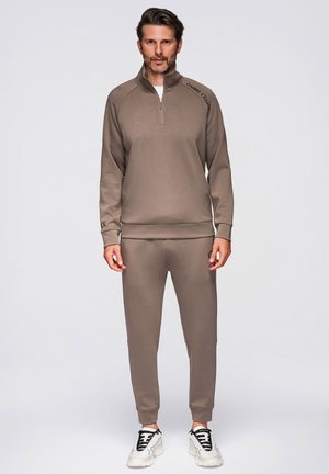 Ein Mann steht frontal zur Kamera, trägt eine braune passende Trainingsjacke und Hose sowie weiße Turnschuhe, vor einem neutralen grauen Hintergrund.