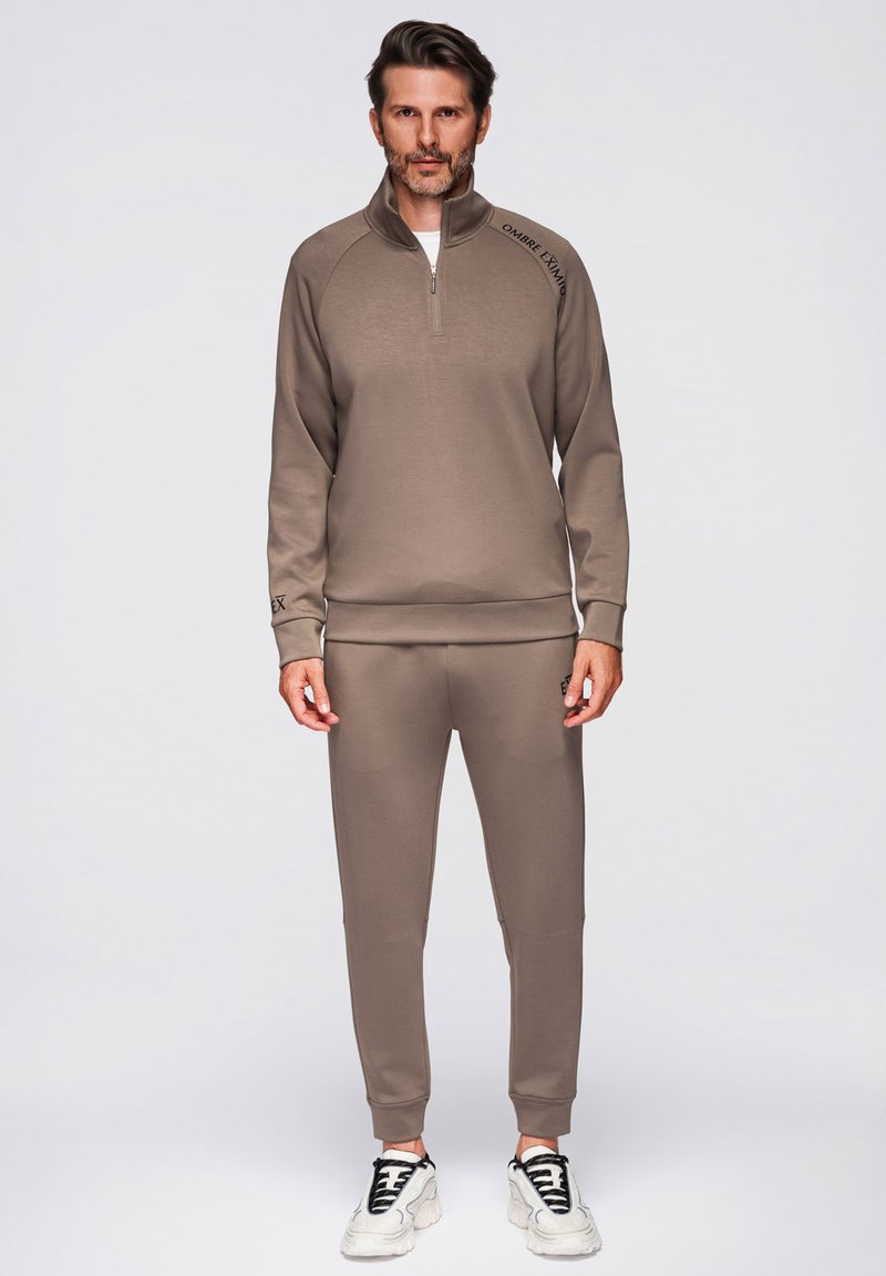 Un homme se tient face à l'avant, portant une veste de survêtement marron assortie et un pantalon, des baskets blanches, sur un fond gris neutre.