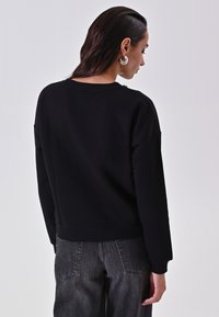 Felpa nera in cotone con vestibilità rilassata, polsini e orlo a coste. Sullo sfondo, un individuo indossa jeans scuri.