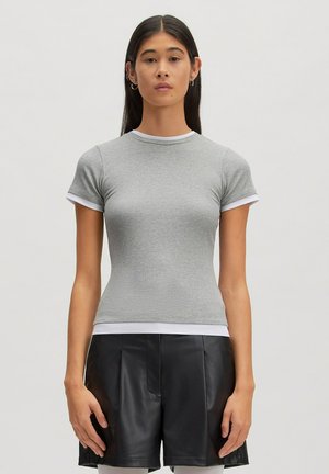 Vrouw met lang donker haar draagt een aansluitend grijs kortmouwshirt met witte bies en zwarte leren korte broek, staand tegen een effen achtergrond.