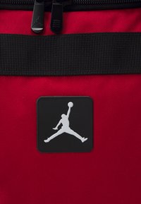 Bolsa de tela roja con cremallera negra, presentan un destacado logo de Jordan en blanco sobre un cuadrado negro. Superficie texturizada con material resistente.
