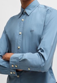 Camisa de botón azul claro fabricada en tela suave, presenta un collar clásico, botones blancos y un pequeño logotipo bordado "BOSS" en el pecho.