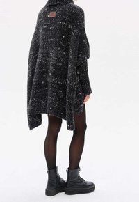 Schwarzer gestrickter Poncho mit grober Textur und hohem Kragen. Verfügt über ein braunes Etikett am Hals und lange, fließende Ärmel. Kombiniert mit schwarzen Stiefeln.