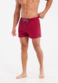 Rode zwembroek met een elastische tailleband en trekkoord, met een zwart logo detail aan de zijkant. Gladde stof, knielange snit.