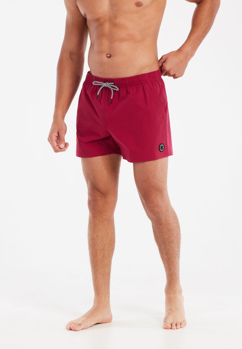 Rode zwembroek met een elastische tailleband en trekkoord, met een zwart logo detail aan de zijkant. Gladde stof, knielange snit.