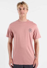 Jeune homme aux cheveux blonds portant un t-shirt rose poudré avec un petit logo circulaire foncé sur la poitrine côté gauche, debout devant un fond blanc.