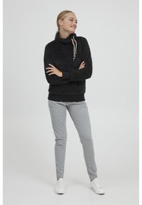 Sort, tekstureret fleece sweater med høj krave, snor lukning, lange ærmer og sort elastik i taljen, sammen med grå sweatpants og hvide sko.