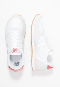 Chaussures de sport blanches avec des accents en daim gris, une semelle en caoutchouc texturée et un détail rouge au talon. Elles présentent un design à lacets et un logo bien visible.