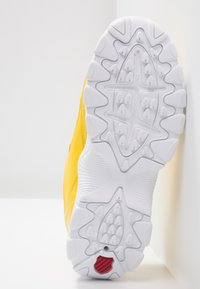 Semelle de chaussure avec bande de roulement blanche, motifs détaillés et petite forme de bouclier rouge près du talon, dessus jaune partiellement visible sur fond blanc.