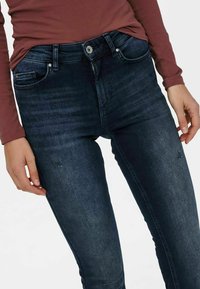 Jean skinny en denim foncé avec une légère décoloration. Comprend des poches avant et un petit patch abîmé près du genou.