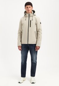 Lichtbeige zip-up jas met een capuchon, voorzien van een diagonale ritszak en verstelbare trekkers. G gedragen met een donkere spijkerbroek en witte sneakers.