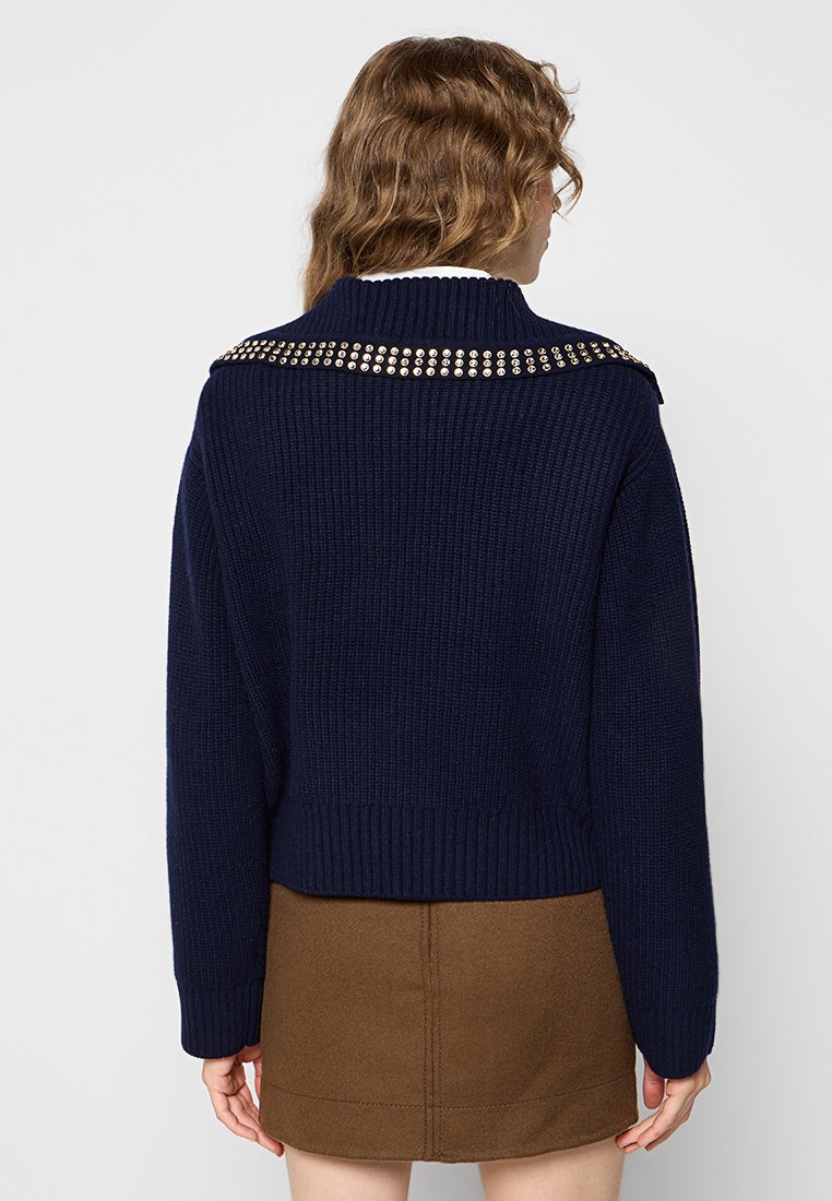 Pull en maille navy avec une texture côtelée et des clous métalliques décoratifs sur les épaules, associé à une jupe marron.