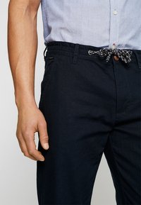 Donkerblauwe katoenen broek met een elastische tailleband, voorzien van een geknoopt trekkoord en een knopdetail aan de voorkant. Soepele textuur, geen patronen.