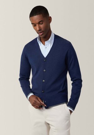 Cárdigan náutico hecho de una suave tela de punto, con cuello en V, seis botones y puños acanalados. Llevado sobre una camisa de rayas azul claro.
