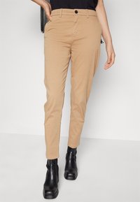Béžové chinos kalhoty se slim fit střihem, s knoflíkovým zapínáním, bočními kapsami a hladkou texturou bavlněného materiálu, doplněné černými kotníkovými botami.
