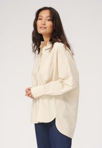 Beige, langærmet skjorte med knapper og spidse krave, to forlommer og rundet kant. Stoffet fremstår let og glat.