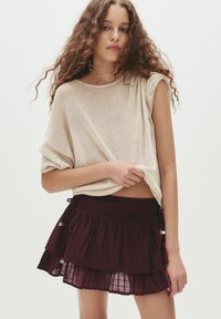 Beige gebreide top met opgerolde mouwen, gecombineerd met een gelaagd bordeauxrood rokje. Het rokje heeft een geraffineerde tailleband en decoratieve zilveren accenten.