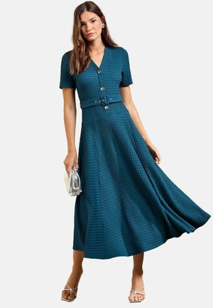 Nainen pukeutuneena struktuurilliseen sinivihreään napilliseen mekkoon, jossa on vyö, pitää hopeista clutch-laukkua ja käyttää valkoisia remmikenkiä.