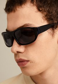 Black sunglasses with large rectangular frames, tinted lenses, and sleek arms, positioned on a model's face with short wavy hair.