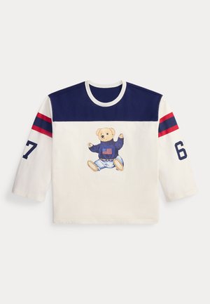 Langärmliges weißes und marineblaues Hemd mit roten Streifen auf den Ärmeln, der Zahl 67 auf den Ärmeln und einem Teddybär, der einen Pullover mit einer amerikanischen Flagge auf der Vorderseite trägt.