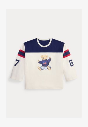 Langärmliges weißes und marineblaues Hemd mit roten Streifen auf den Ärmeln, der Zahl 67 auf den Ärmeln und einem Teddybär, der einen Pullover mit einer amerikanischen Flagge auf der Vorderseite trägt.