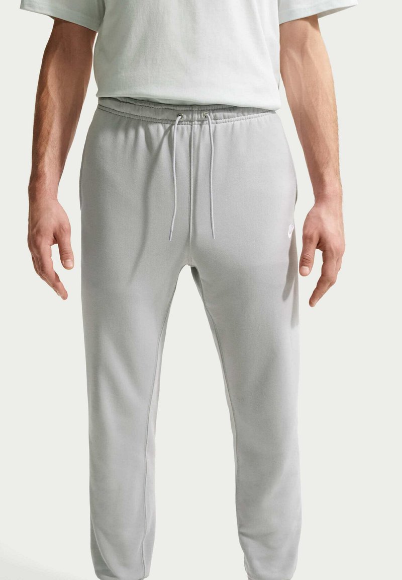 Hombre vistiendo pantalones deportivos gris claro con cintura de cordón y logo blanco en el muslo izquierdo, combinado con una camiseta blanca.