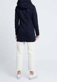 Donkerblauwe gewatteerde jas met een lange snit, lichtgewicht stof en gestikte details. Gecombineerd met witte cropped broek en witte sneakers.