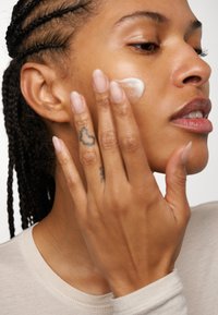 Ihonhoitotuotetta levitetään mallin poskelle; malli on punottu hiuksista, käyttää neutraalin väristä paitaa ja luonnolliset kynnet, joihin on tatuoitu sormiin.