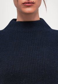 Gros plan sur le bas du visage et le cou portant un pull en maille côtelée bleu foncé avec un petit grain de beauté au-dessus de la lèvre supérieure.