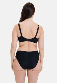 Mujer con el pelo largo y oscuro, que lleva un sujetador negro y ropa interior negra de talle alto, vista desde atrás con un fondo liso.