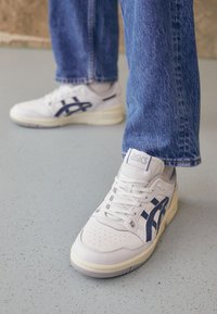 Baskets blanches avec une surface texturée, ornées d'accents bleu marine et d'un logo, bout arrondi et semelle rembourrée, associées à un jean en denim.